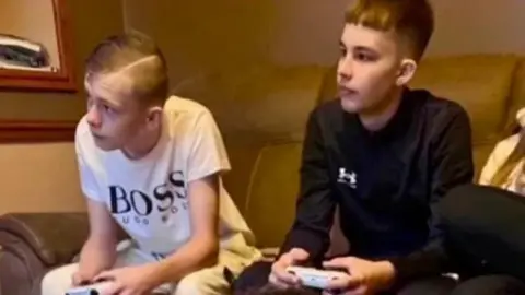 Mason Rist and Max Dixon are shown sitting on a sofa playing a video game together. Both hold controllers and look towards a screen out of shot, with their forearms leaning on their knees. Mason wears a white T-shirt while Max has a long-sleeved black top on.