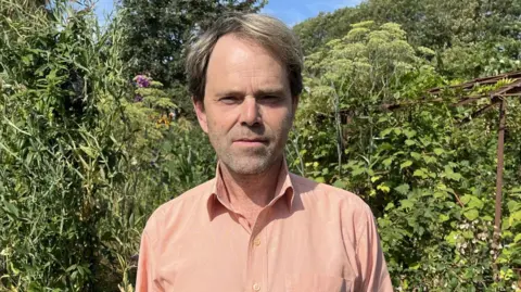 Qays Najm/BBC Environmental campaigner Professor Rupert Read in his Norfolk garden. He is looking directly at the camera and there is a lot of green foliage behind him. 