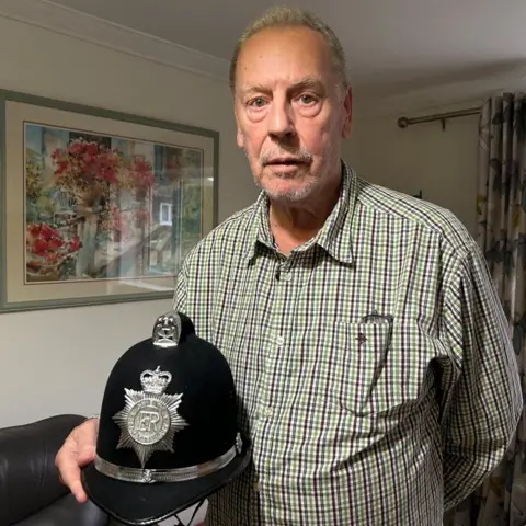 Mark is standing indoors holding a traditional British police helmet featuring a silver badge with the letters “ER” and a crown emblem. The individual is wearing a checked shirt, and a pair of glasses is tucked into the shirt pocket. Behind them, the room has cream-colored walls, patterned curtains, and a framed painting of a floral scene hanging on the wall.