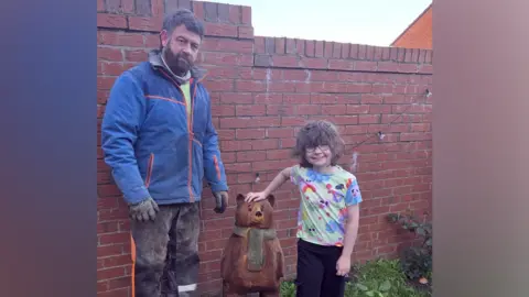 A young girl with short wavy brown hair is wearing glasses and a multi-coloured T-shirt and has her hand on top of a small brown wooden bear with green scarf sculpture in a garden. A man stands on the other side of the bear. He has brown hair and a beard, and is wearing a blue waterproof jacket and black trousers with sawdust on them, as well as black gloves.