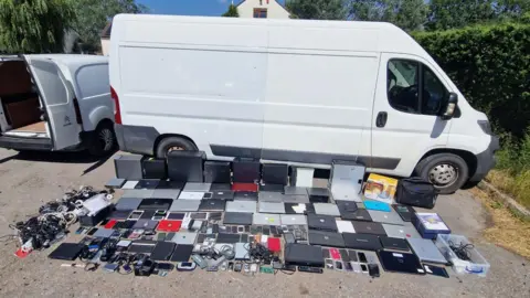A white van parked in a car park beside a hedge. On the ground beside the van there is a large haul of digital devices that have been laid out on the concrete, including laptops, phones, tablets, camera and charging cords. 