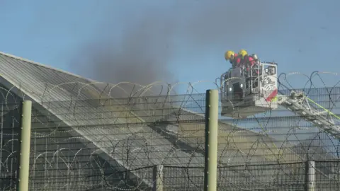 Firefighters in the cage of an aerial ladder platform. There appear to be two of them. They are looking at a roof from which billowing black smoke is coming. The shot has been taken through a barbed wire topped wire fence at Highpoint Prison.