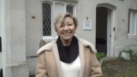 Local Democracy Reporting Service/Oliver Leader de Saxe A blonde woman wearing a beige-colour jacket and a white top and black snood. She is standing in front of a property.