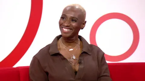 Ada Osigwe smiles as she does a studio interview. She wears a brown shirt open at the collar with chains above her chest scar.