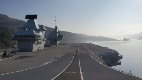 DAVE ROBSON A general view of the flight deck on board the HMS Prince of Wales. The runway is grey with white lines on it. It is slightly curved in the foreground of the image leading towards the loch and mountains in the background. Two grey towers are on the left of the image.