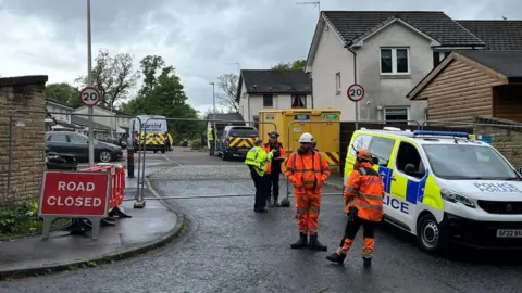 Police officers and workers wearing hi-vis stand outside a blocked-off road 