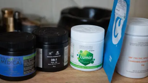 White and black plastic medical containers lined up on a wooden counter top. Some have blue and green coloured labels stating the containers carry medical cannabis.