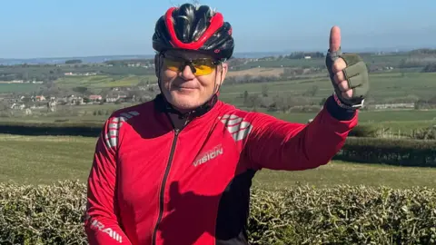 Freddie Peart, wearing a red cycling top, cycling gloves, a helmet and sunglasses, gives a thumbs up as he stands in countryside.