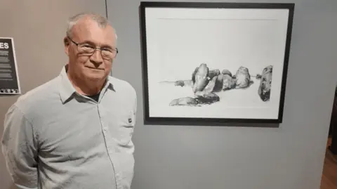 Andrew Makellar is wearing a grey top. He is standing by his black-and-white drawing of a stone site on the Isle of Man. 