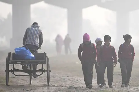 AFP via Getty Images Four children in school uniforms walk through a field on a smoggy winter morning in New Delhi on 17 December. Beside them, a man riding a cycle cart