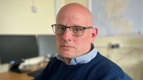 David Murray, a bald clean shaven man with glasses turns to the camera while sitting at a desk with two screens on it. A map is on the wall behind him. He wears a blue checked shirt and a navy jumper.