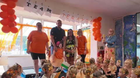 Two women in orange t-shirts with Teraysa, who is in an orange t-shirt and pink helmet, with a man wearing a black t shirt and jeans, and a woman in a floral jumpsuit. A class of small children is also present.