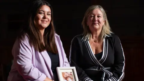PA Carley Barret has long dark hair and is smiling. She is holding a photo of her sister with her baby. Sitting next to her is her mother Alison Madgin who is wearing a black dress with white piping and is also smiling. The background of the photo is black. 