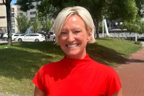Ruth stands and smiles at the camera. She is wearing a red dress and has short blonde hair. It is a head and shoulders shot of her. Behind her is a grass verge and a path, with cars to the left. 