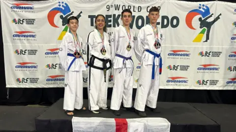 Cambridge Taekwon-Do Three boys and a woman, all in white martial arts outfits, stand on a podium, upon which a St George's flag has been draped. Behind is a backdrop that says "9th World Cup Taekwon-do" and features sponsors' logos.