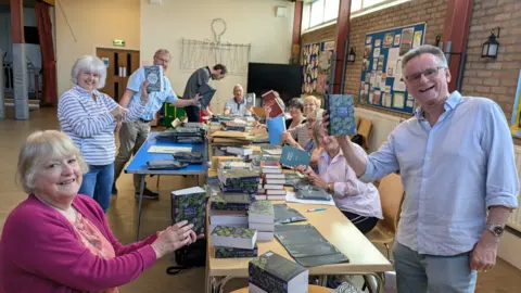 St Thomas Church A group of ten men and women sit and stand around long tables stacked with books inside a hall. Several of them are smiling and holding the books up. Envelopes can be seen on the table. Noticeboards can be seen on the wall to the right.