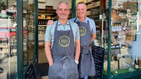 BBC/Angela Ferguson A man with short grey hair and a beard stands next to another man with short grey hair. Both are smiling and the one on the right has his hand on the shoulder of the other man. They are wearing denim aprons with yellow Birkdale Cheese Co logos on them. A window display on the right includes what looks like a bottle of sparkling wine next to a glass, some bags with the shop logo on them and a variety of products including what appear to be drinks, dressings and chutneys.