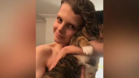 Ellen Roome A young teenage boy with dark blond hair smiles as he cuddles into a tabby cat.