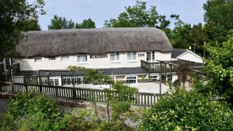 The exterior of the pub. It has large black letters that read: THE BLAGDON INN under a long thatched roof. The building has two storeys and there is a courtyard area surrounded by wooden fencing. There is mature planting and trees surrounding the property.