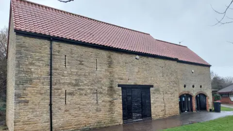 Google A barn with a red roof and three black gates placed along the length of building with some greenery in front of it.