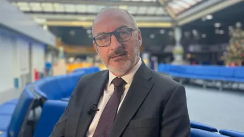 BBC Christophe Mathieu - a man wearing black rectangular glasses, a grey suit with a white shirt and purple tie, sitting on blue chairs in the harbour ferry terminal.