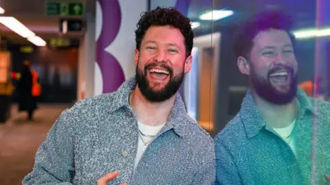 A man stands against a window in a radio studio with a broad smile on his face. He has curly brown hair, a brown beard and wears a chunky blue cardigan and white shirt. His reflection is clear in the window.
