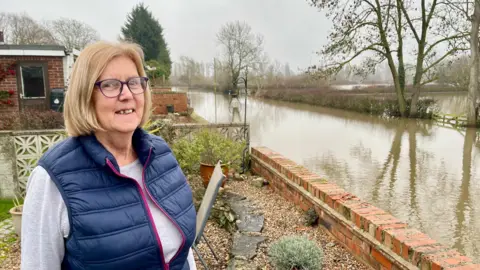A woman stands in her gravelled garden. At the end of her garden there is has a wall which drops down to the road. That road is completely flooded with brown water, as are the fields on the other side of the road. Hedgerows, a gate and trees are partially underwater. The woman has short blonde hair, glasses, a blue body warmer and long-sleeved grey top. There are plants in pots and a chair in her garden. Next-door's garden is separated by a grey stone wall.