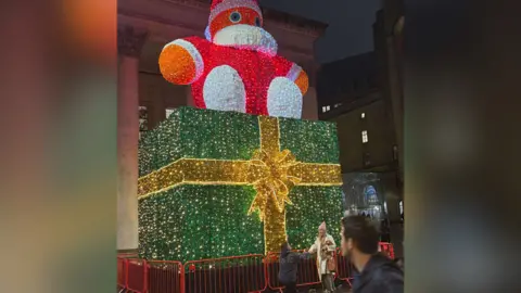Charlotte Busby Different angle - man is down on one knee in front of a Christmas light display in Manchester's St Peter's Square. The lights show a big green present wrapped with a bow and a big orange Santa Claus positioned on top. The man has brown hair and wears a raincoat the woman, blonde has long hair, wears a pink wooly hat and a white coat.