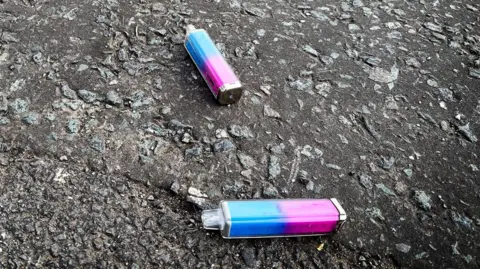 Two single-use vapes on a concrete path or road. They are plastic on the outside, and blue and pink in colour.