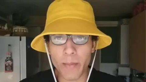 YouTube Nicholas Prosper, mengenakan kacamata dan topi ember kuning dengan tali putih, berbicara di kamera dalam video buatan sendiri di dapur.