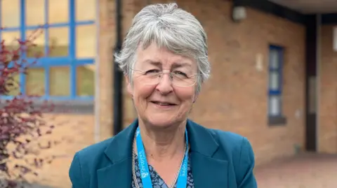 A woman with grey hair in a blue jacket smiles at the camera. She is wearing frameless glasses and a blue lanyard around her neck. She is standing outside a building. A tree can be seen to her left.