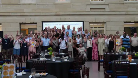 Wiltshire Council A large group of people posing for the camera with several of them holding their hands in the air. There is a stone wall behind them with windows and a large screen in front. In the foreground are several round tables with the remains of food and drinks on them.