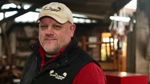 Martin Giles/BBC Andy Warnes is smiling at the camera. He is wearing a company branded white hat and black gilet with a red shirt underneath. He is standing in a store with blurred furniture items behind him.