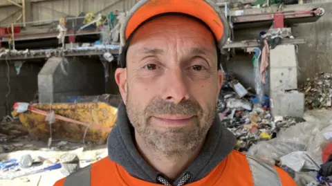 A man with dark stubble, who is wearing an orange cap and hi-vis jacket stands in front of piles of scrap in a warehouse.