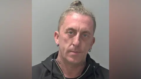 A police custody image of a man with a blond top knot, a black hoodie and small silver hoop earrings. He has some tattoos on his neck.