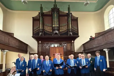 A male voice choir dressed in blue blazers and blue ties and holding choral hymn books sing in front of a large ornate pipe organ. 