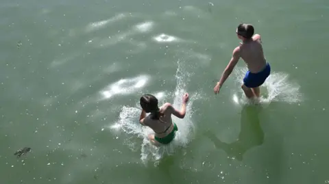 Two children jump into a body of water wearing swimming costumes