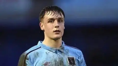 Family handout Alexander Birnie pictured while playing football. He's standing with mud flecks on his face and has short dark hair. He is wearing a light blue football shirt covered in mud marks.