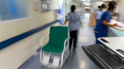 PA Media A large green and white wheelchair being moved at speed down a hospital corridor by a member of staff while two others stand nearby looking at notes. There is a black computer keyboard in the foreground. 