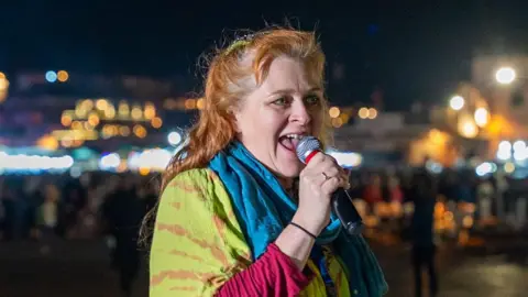 Justine de Mierra is pictured speaking into a microphone. She has long red hair and wears a green and red top with red sleeves as well as a blue scarf.