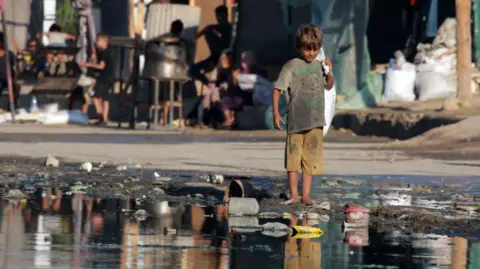 Getty Images A Palestinian boy stands barefoot near stagnant wastewater in Deir el-Balah, in the central Gaza Strip (19 July 2024)