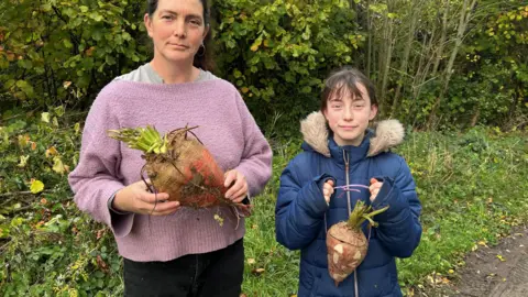 BBC Dawn Qince, with brown hair and wearing a purple jumper, is holding a Mangelwurzel to carve and and her daughter Cherry, wearing a blue coat, is holding a carved lantern. They are both stood outside with bushes in the background.