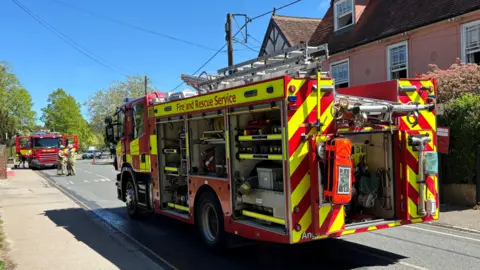 A fire engine parked in a residential road, pictured from behind. There is an old pink house the other side of the vehicle. There is a blue sky, and another fire engine in the background.