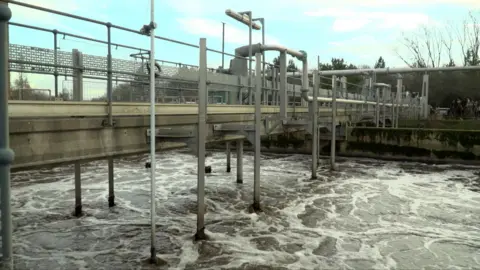 Ben Schofield/BBC A footbridge over a swirling body of water at Bedford Water Recycling Centre. The bridge is held up by metal stilts that are sinking into the water, the surface of which is flecked with white as currents battle underneath.