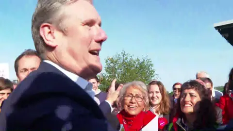A still of TV footage of Keir Starmer talking to a group of Labour campaigners in an outdoor area. It is a sunny day and there are trees visible in the background. 