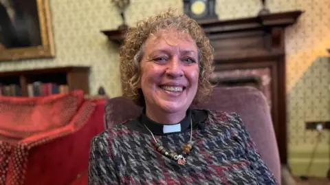 A woman with curly brown hair smiles as she sits in an armchair in a large sitting room. She is wearing a black, grey and red patterned pullover, a black shirt, a white dog collar and a beaded necklace. The room is dominated by a large dark-wood fireplace and decorated with cream wallpaper with a cross pattern. A red sofa stands to the left in front of a dark-wood bookcase and a brass-framed painting.