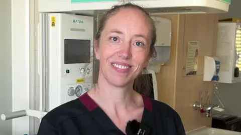 A woman with blonde hair is pictured smiling at the camera in a hospital ward. 