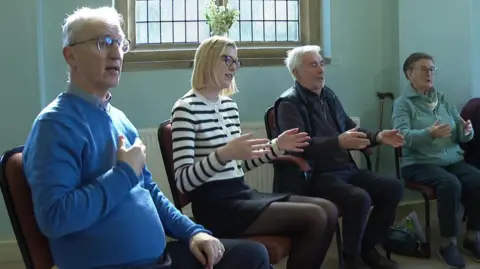 BBC Two men and two women sitting in a row on chairs. All four have their hands raised in front of them as part of a breathing exercise linked to singing.