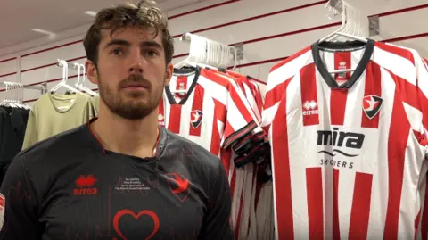 A photo showing Cheltenham Town defender Sam Stubbs stood in front of a row of football shirts. He is wearing a black football shirt