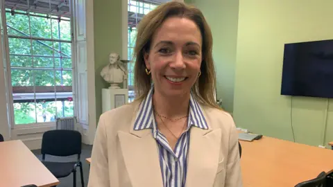 Dr Rebecca Foljambe is standing in a room with green walls. She is wearing a shuit jacket and a blue and white shirt. She is smiling.
